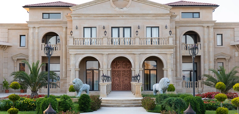 Elegante villa met beige stenen gevel, grote gebogen ingang, balkon en weelderige tuinen. Flankeren door leeuwenbeelden, straalt grandeur en luxe uit.